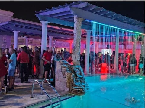 The pool deck and pergola early in a TampArotic hotel-takeover evening in Tampa Bay, with guests lit by neon at the water's edge.